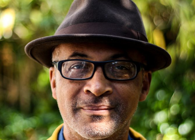 An image of the Black writer Colin Grant taken outside. He is wearing glasses and is sporting a brown hat, blue shirt and yellow jacket.