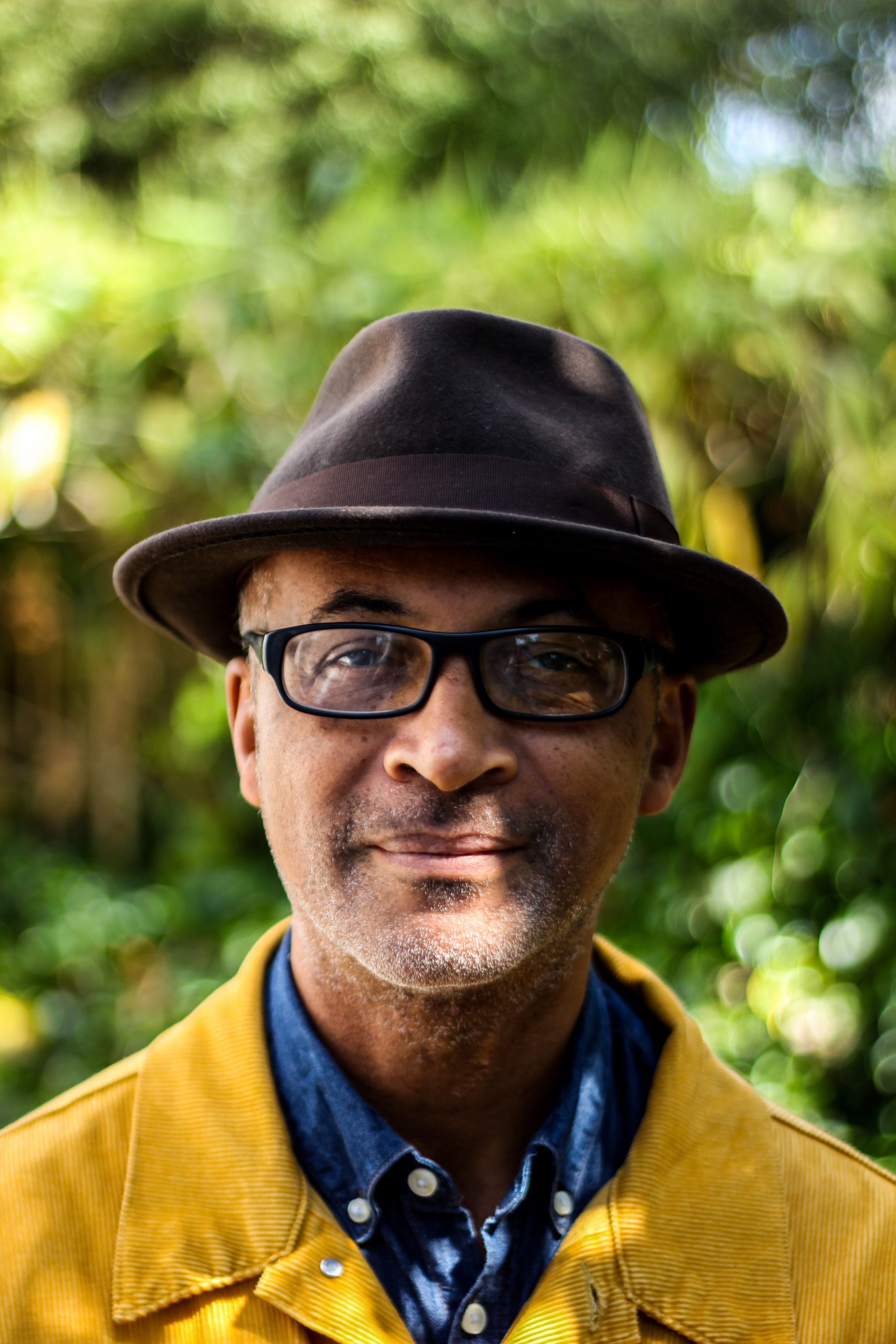 An image of the Black writer Colin Grant taken outside. He is wearing glasses and is sporting a brown hat, blue shirt and yellow jacket.