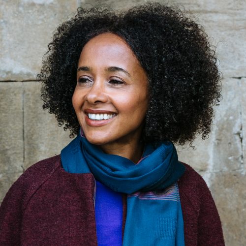 A portrait of Black novelist and journalist Diana Evans. She has shoulder length hair and is wearing a maroon jacket, blue shirt and blue scarf.