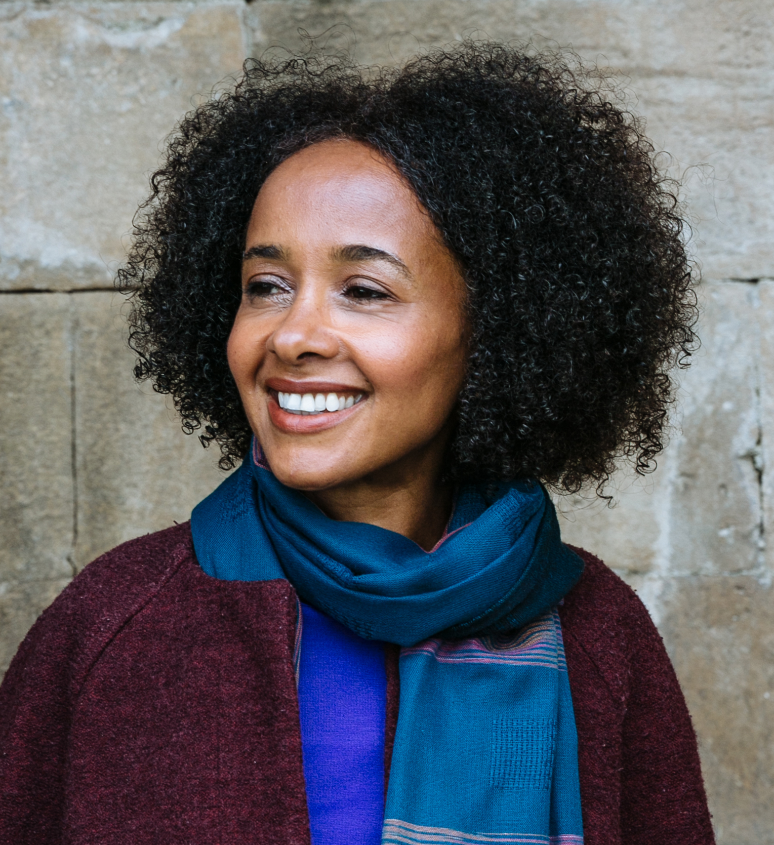 A portrait of Black novelist and journalist Diana Evans. She has shoulder length hair and is wearing a maroon jacket, blue shirt and blue scarf.