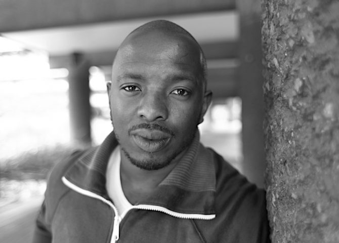 A portrait of Black poet Nick Makoha. He is pictured outside leaning against a wall. He's wearing a white t-shirt and tracksuit top.