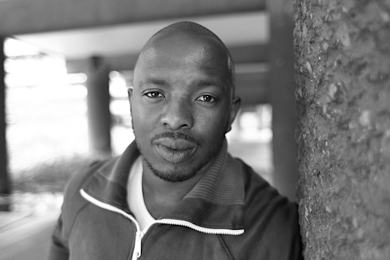 A portrait of Black poet Nick Makoha. He is pictured outside leaning against a wall. He's wearing a white t-shirt and tracksuit top.