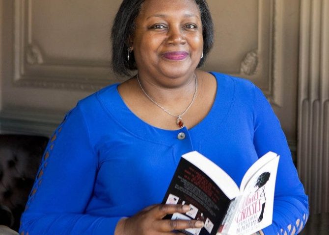 An image of Malorie Blackman. She has chin length black hair and is wearing a round necked long sleeved top and a necklace. She's holding a copy of Noughts and Crosses