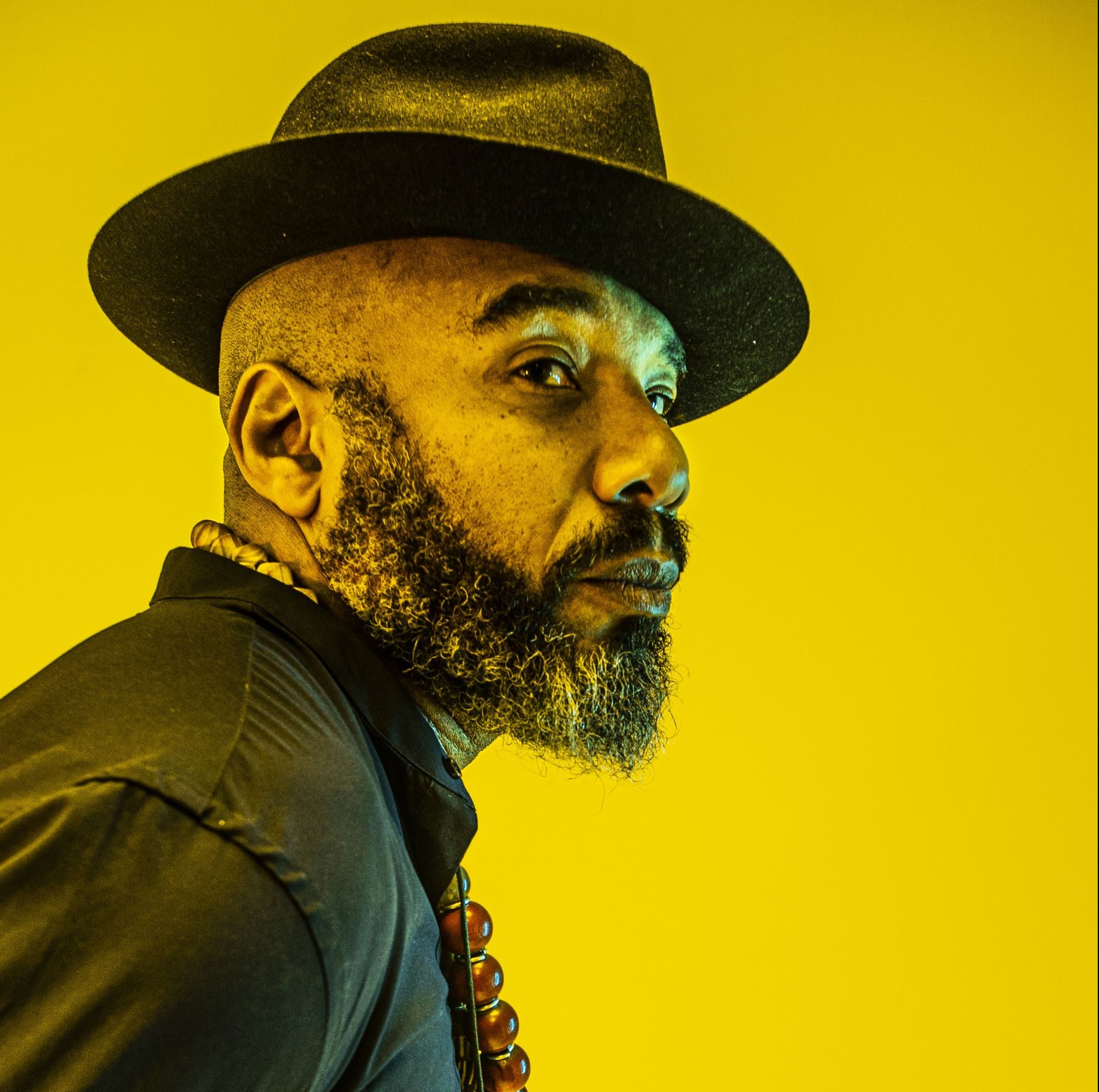 An image of Caribbean writer Anthony Joseph. He is wearing a black trilby, black shirt and is bearded. This image is a side profile and features a yellow background.