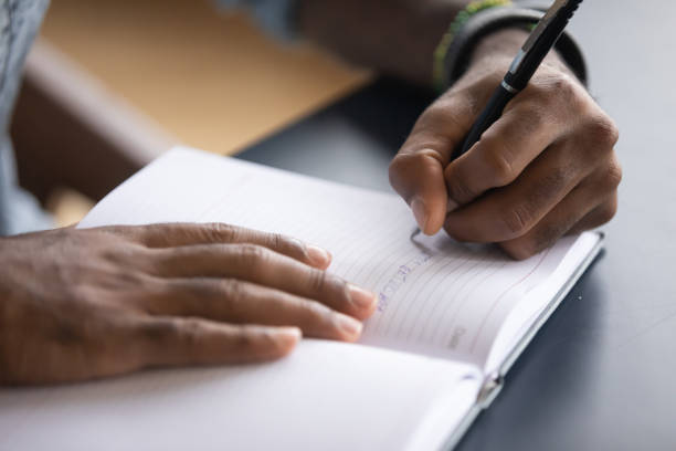 A person of colour sits at a desk. We can see his or her hands holding a pen in the left hand writing notes into a book which is lying on the desk in front of them.